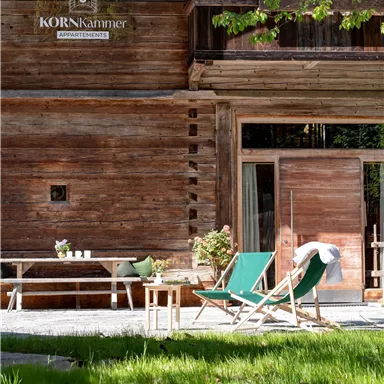 A cozy wooden terrace with two lounge chairs on green grass. In the background, there is a rustic wooden house with the logo "KORNkammer".