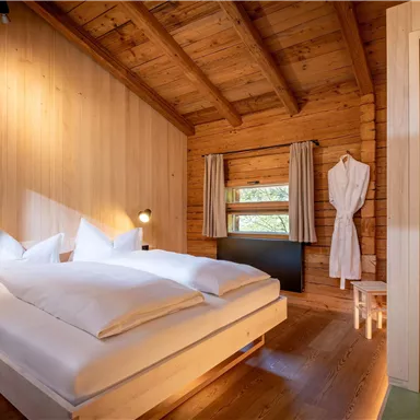 A cozy bedroom in a wooden house with a double bed and soft lighting. In the background, a bathroom is visible, and the walls are made of light wood.