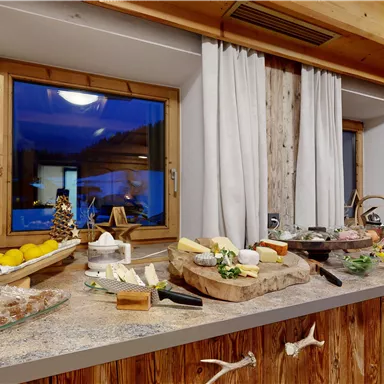 A buffet with various types of cheese, cold cuts, and fresh fruit. The room has large windows with a view outside and a cozy wooden structure.