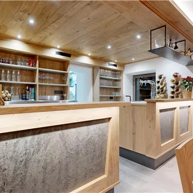 A modern bar with a wooden counter area and warm lighting accents. In the background, there are shelves with glasses and decorations.