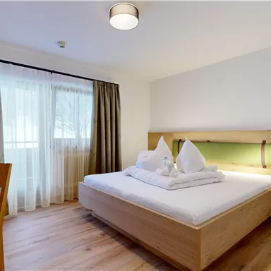 A bright hotel room with a double bed and a wooden desk. In front of the window are long curtains and the floor is made of wood.