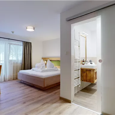 A modern hotel room with a large bedroom and a bathroom. The space is bright and friendly decorated, with wooden furniture and a window.