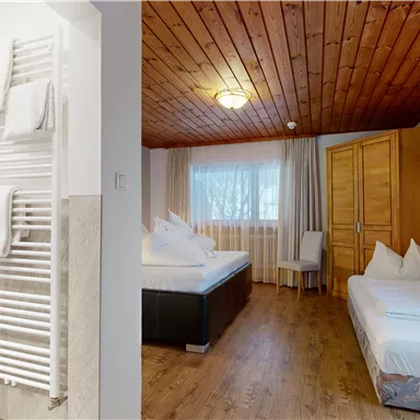 A cozy room with a wooden ceiling, a double bed, and a single bed. On the left side, there is a modern bathroom with a towel radiator.