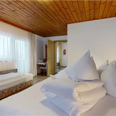 A cozy room with wooden floor and bright wooden ceiling. There is a double bed with many pillows and a single bed on the side.