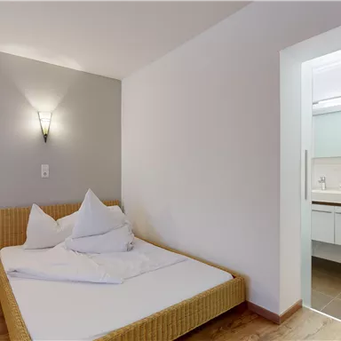 A simple bedroom with a bed and two pillows. In the background, a bathroom is visible.