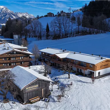 A picturesque snowy landscape with several wooden houses in the foreground. In the background, the snow-covered mountains and a blue sky can be seen.