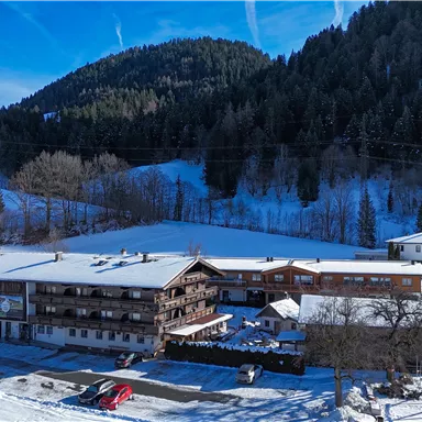 A rustic hotel in a snow-covered landscape. In the background, forested mountains and a blue sky can be seen.