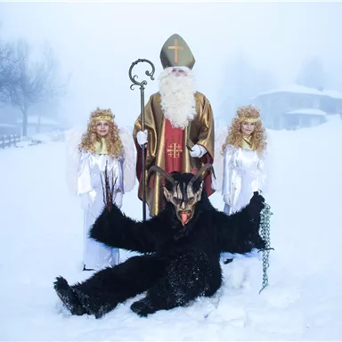 Eine winterliche Szene mit einem heiligen Nikolaus, der von zwei Engeln begleitet wird. Vor ihnen sitzt eine teuflische Gestalt im Schnee.