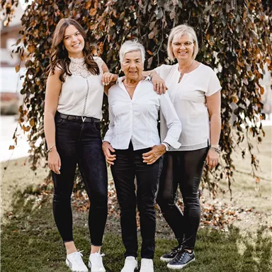 Drei Frauen stehen lächelnd vor einem Baum. Sie tragen moderne, elegante Kleidung in Schwarz und Weiß.