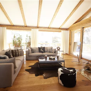Ein modernes Wohnzimmer mit hellen Wänden und einer Holzdecke. Es gibt bequeme Sofas, einen Couchtisch und große Fenster, die viel Licht hereinlassen.