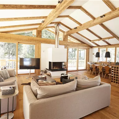 Ein modernes Wohnzimmer mit Holzdecken und großen Fenstern. Es gibt gemütliche Sofas, einen Esstisch und einen Kamin.