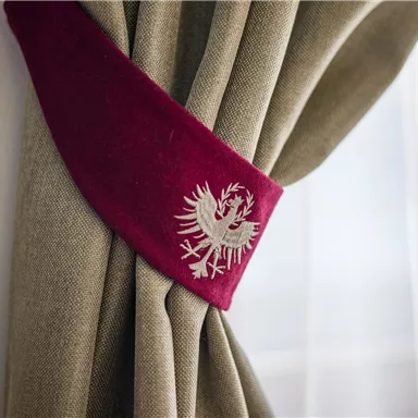 Ein Vorhang mit einer dekorativen Halterung in Burgunderrot. Auf der Halterung ist ein silbernes Wappen mit einem Adler abgebildet.