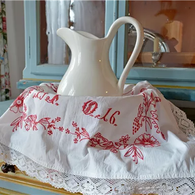 Eine elegante Kanne aus Keramik steht auf einem liebevoll dekorierten Tuch. Im Hintergrund ist ein Holzschrank mit Blumenmuster zu sehen.