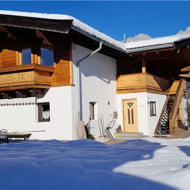 A beautiful, modern house in the snow with wooden elements and a balcony. In the background, the mountains are visible and the sky is clear blue.