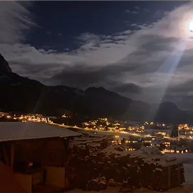 A nocturnal mountain landscape with snow-covered peaks and a bright moon. The lights of a village twinkle in the valley.