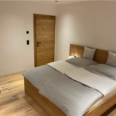 Ein modernes Schlafzimmer mit einem großen Holzbett und zwei Kissen. Der Boden ist aus Holz, und es gibt einen Schreibtisch sowie eine Holztür.