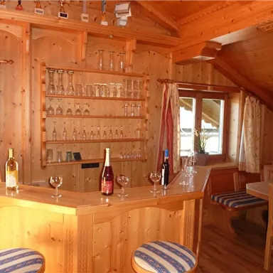 A cozy wooden bar with elegant glasses and several bottles of wine. In the background, there is a window and a seating area with comfortable chairs.