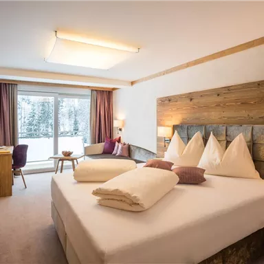 Ein modernes Hotelzimmer mit einem großen Bett und warmen Holzelementen. Große Fenster bieten einen Blick auf eine winterliche Landschaft.
