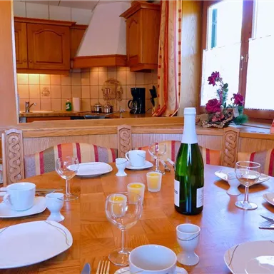An inviting kitchen scene with a set table featuring dishes and glasses. In the background, wooden furniture and a well-equipped kitchen unit can be seen.