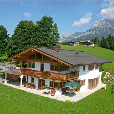 Ein schönes, modernes Haus inmitten einer grünen Wiese. Im Hintergrund sind majestätische Berge und ein blauer Himmel zu sehen.