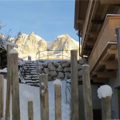 Eine Schneelandschaft mit verschneiten Hütten und einem Holzzaun. Im Hintergrund sind majestätische Berge unter klarem Himmel zu sehen.
