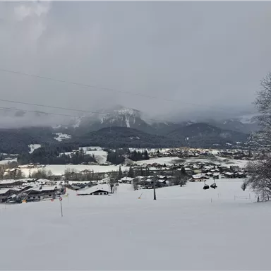 A snowy landscape with small houses and mountains in the background. The sky is overcast and conveys a calm winter atmosphere.