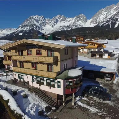 A cozy hotel in the mountains, surrounded by snow-covered landscapes. The majestic mountains rise in the background under a clear blue sky.