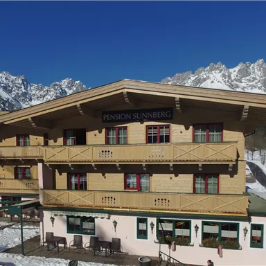 A cozy guesthouse in the snow with a magnificent mountain view. The facade is designed in a traditional style, surrounded by a wintry landscape.