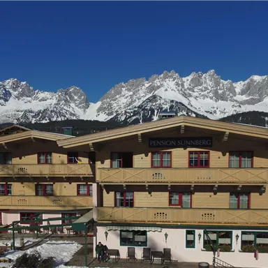 A cozy chalet in the mountains with snow-covered peaks in the background. Clear blue skies and a tranquil setting.