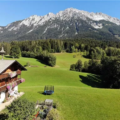 Ein malerisches Alpenpanorama mit schneebedeckten Bergen im Hintergrund. Im Vordergrund steht ein traditionelles Haus umgeben von grünen Wiesen und Bäumen.