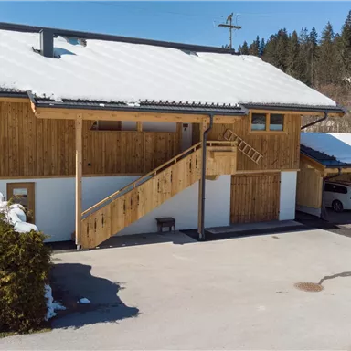 Ein modernes Gebäude mit Holzverkleidung und einem schneebedeckten Dach. Auf der rechten Seite befinden sich Parkplätze und ein Weg mit einer Treppe.