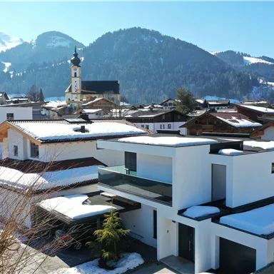 Ein modernes Haus in einer schneebedeckten Landschaft mit Bergen im Hintergrund. Im Vordergrund sind traditionelle und zeitgenössische Architektur zu sehen.