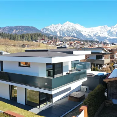 Ein modernes, zweigeschossiges Haus mit großen Fenstern und Balkon. Im Hintergrund sind schneebedeckte Berge und eine idyllische Landschaft zu sehen.