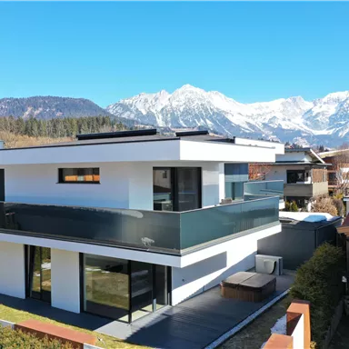 Ein modernes, weißes Einfamilienhaus mit großen Fenstern und einem Balkon. Im Hintergrund sind beeindruckende Berge und ein klarer blauer Himmel zu sehen.