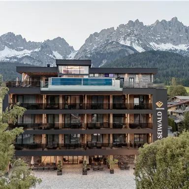 Ein modernes Hotelgebäude mit mehreren Etagen und großen Fensterfronten. Im Hintergrund erheben sich beeindruckende Berge unter einem klaren Himmel.
