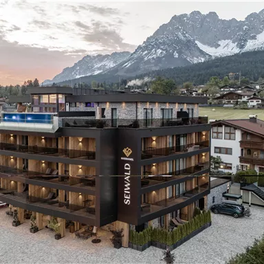 Ein modernes Hotel mit mehreren Stockwerken liegt vor einer malerischen Berglandschaft. Im Vordergrund sind parkende Autos und eine ruhige Umgebung zu sehen.