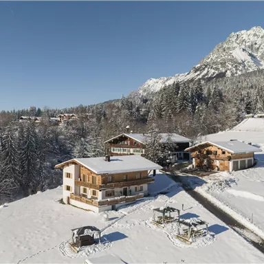 Eine malerische Winterlandschaft mit schneebedeckten Hügeln und traditionellen Holzhäusern. Im Hintergrund ragen majestätische Berge unter einem klaren blauen Himmel empor.