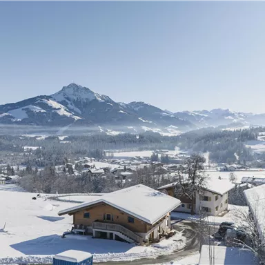Eine malerische Winterlandschaft mit schneebedeckten Hügeln und Bergen im Hintergrund. Im Vordergrund sind traditionelle alpine Häuser zu sehen.