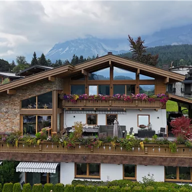 Ein modernes Haus in den Bergen mit Holzverkleidung und bunten Blumen. Im Hintergrund sind majestätische Berge und eine bewölkte Himmel.