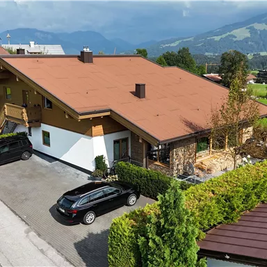 Ein modernes Haus mit einem roten Dach in einer ruhigen Wohngegend. Umgeben von grüner Landschaft und Bäumen, stehen mehrere Autos auf der Einfahrt.