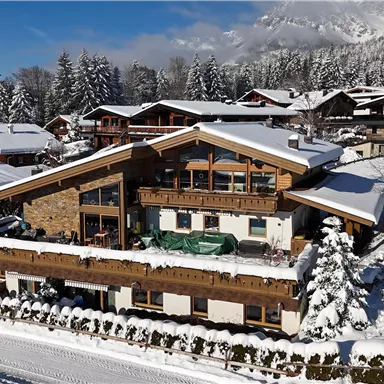 A cozy chalet in the snow with many snow-covered trees all around. The environment is quiet and idyllic with a clear mountain backdrop.