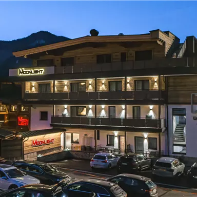 A modern hotel building at night, surrounded by mountains. Parking spaces are visible directly in front.