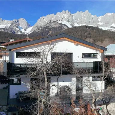 Ein modernes Haus in den Bergen, umgeben von schneebedeckten Landschaften. Im Hintergrund sind majestätische Berggipfel und ein klarer blauer Himmel zu sehen.