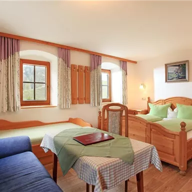 Ein gemütliches Zimmer mit zwei Betten und einem blauen Sofa. Die Fenster lassen viel Licht herein und die Einrichtung ist Holz- und Naturfärbungen.