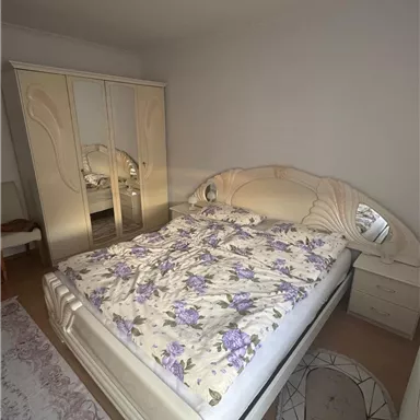 A bright bedroom with a large bed and white bedding adorned with a purple floral pattern. In the background, there is a large wardrobe with a mirror.