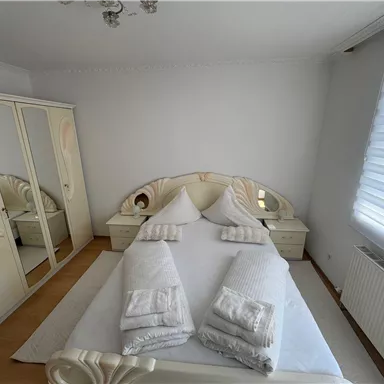 Ein helles Schlafzimmer mit einem großen Bett und beigen Wänden. Es gibt einen Spiegel und einen großen Kleiderschrank im Raum.