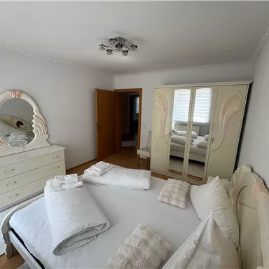 Ein helles Schlafzimmer mit einem großen Bett und einer eleganten Kommode. Der Raum ist schlicht und modern eingerichtet, mit einem großen Spiegel und einem geräumigen Kleiderschrank.