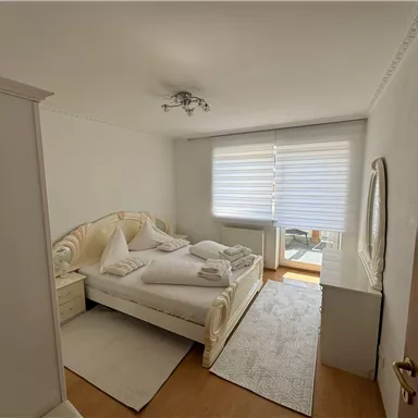 Ein helles Schlafzimmer mit einem großen Bett und weißer Bettwäsche. Es gibt einen großen Fenster mit Jalousien und einen Spiegel an der Wand.