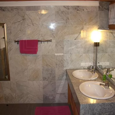 Ein modernes Badezimmer mit zwei Waschbecken und großem Spiegel. Die Wände sind mit grauen Fliesen gestaltet, und ein pinkes Handtuch hängt an der Wand.