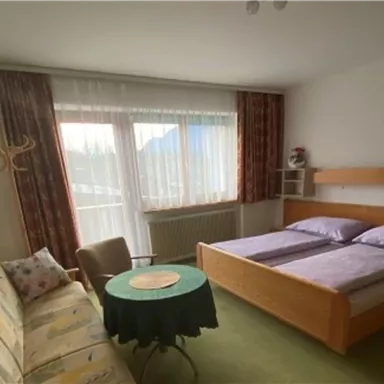 Ein einfaches Zimmer mit einem Doppelbett, einer Couch und einem kleinen Tisch. Lichtdurchflutete Fenster sorgen für eine angenehme Atmosphäre.